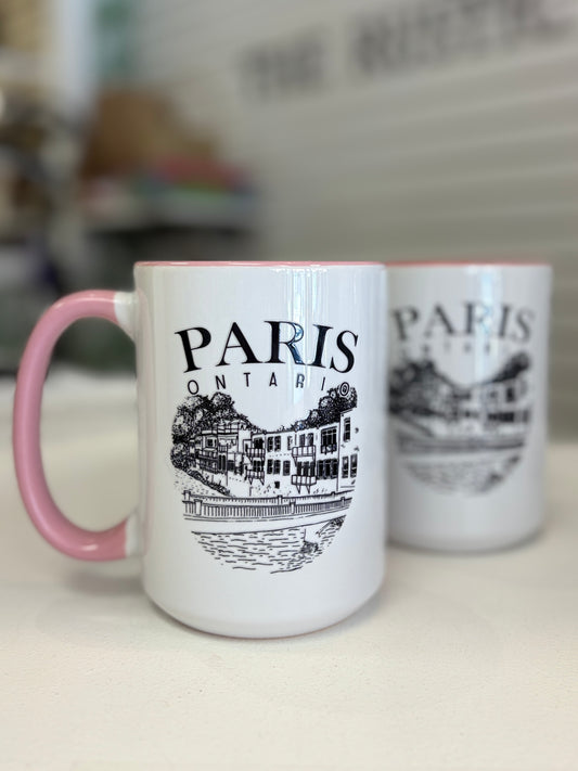 A white ceramic coffee mug with a pink handle and interior, featuring a black line art illustration of Paris, Ontario, with the text 'PARIS ONTARIO' printed on the front. A second identical mug is visible in the background.
